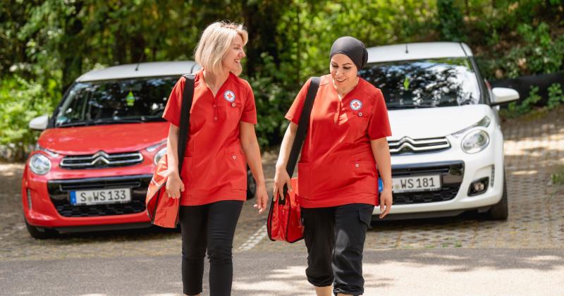 Zwei Frauen unterwegs mit Umhängetaschen. Hintergrund: zwei Autos in rot und weiß